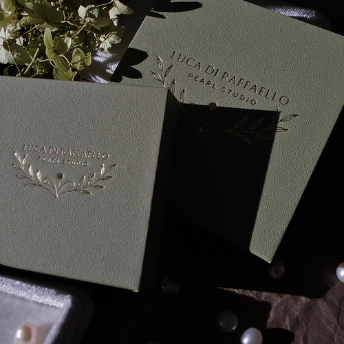 Two embossed light sage green jewelry boxes with gold foil leaf designs, branded 'LUCA DI RAFFAELLO PEARL STUDIO', arranged on a dark surface with scattered pearls and a small plant in the background.