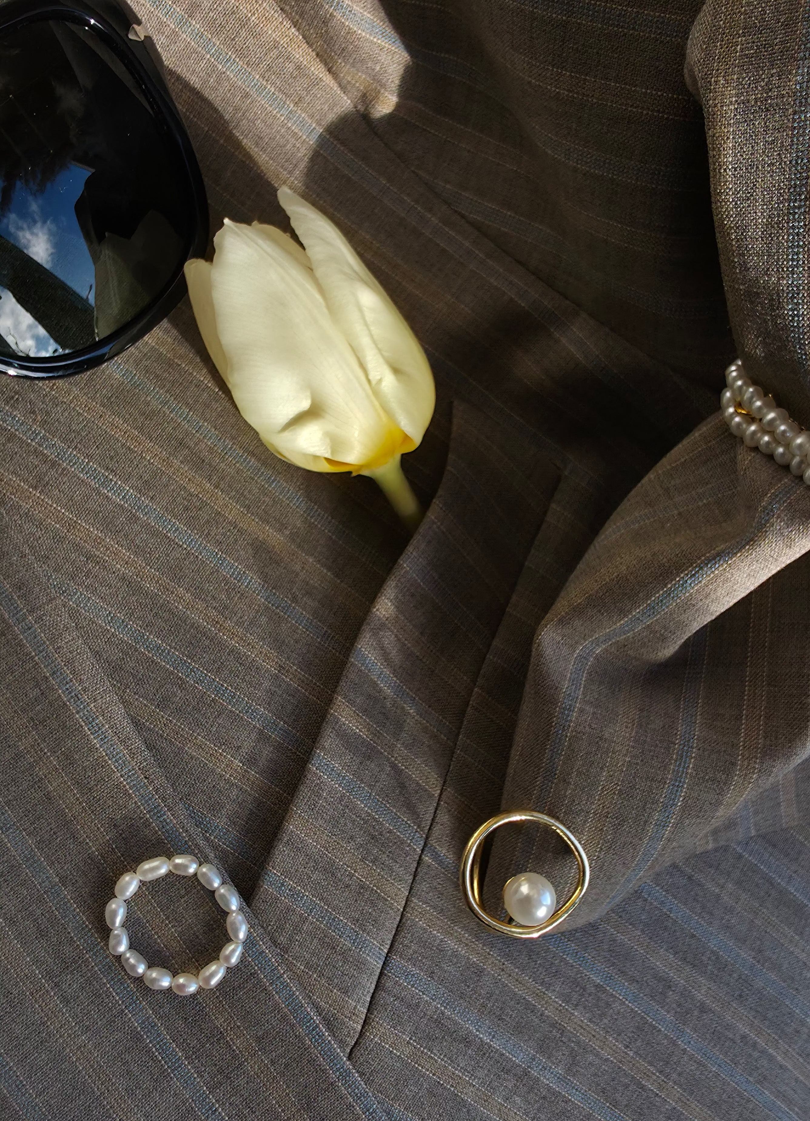 Close-up of a gray suit jacket with a yellow tulip, black sunglasses, and a silver ring on a textured surface.