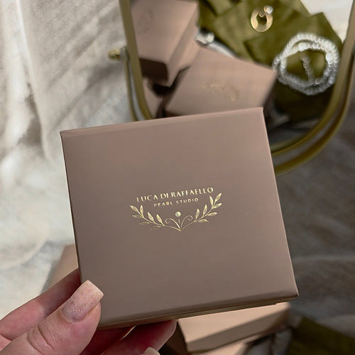 A hand holds a square taupe jewelry box with gold foil branding that reads 'LEA KIM PAMPRELLI' and a leaf motif. The background shows a styled flat lay of olive green velvet jewelry pouches and white pearl necklaces and rings.