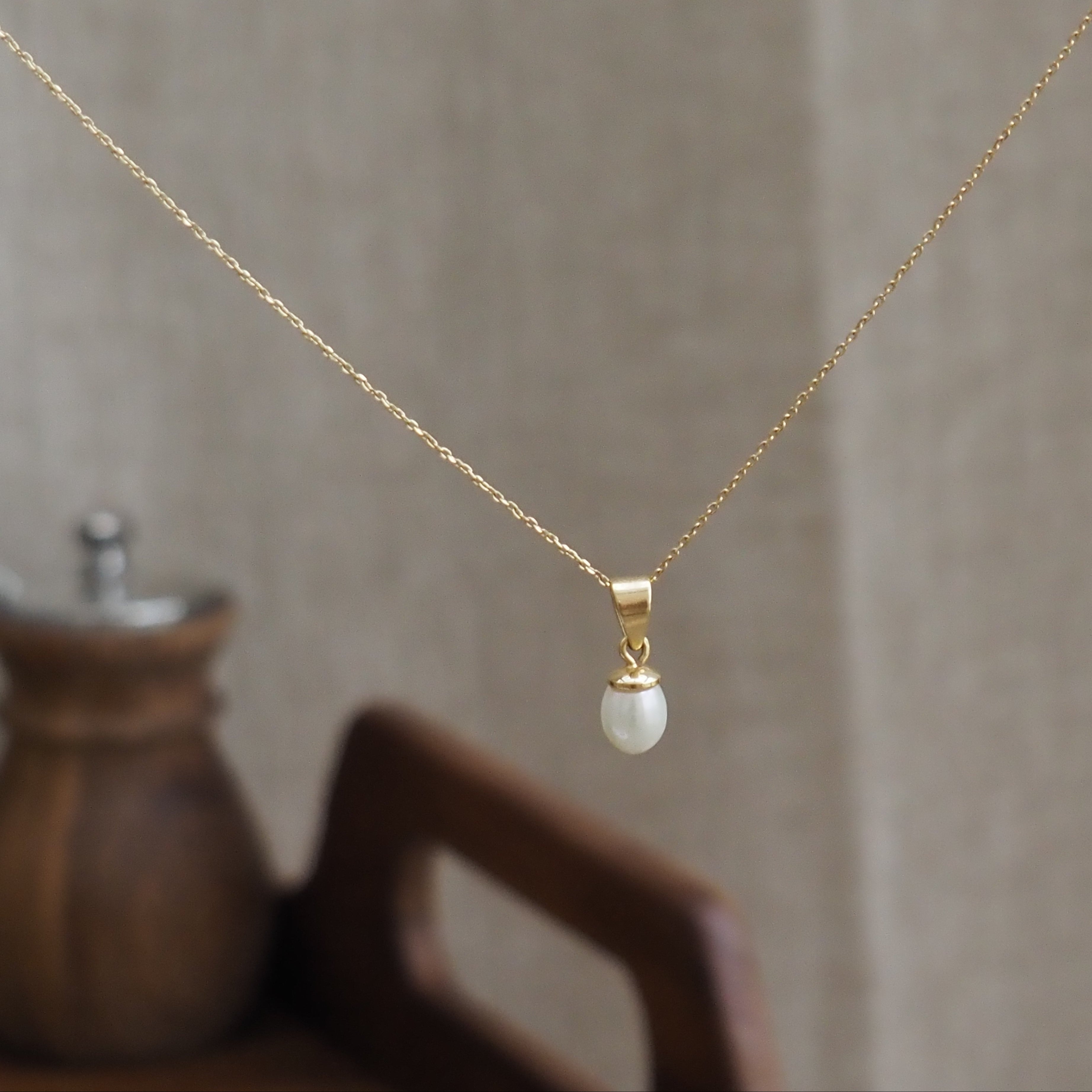 A delicate gold chain necklace with a small gold cap and a single natural pearl pendant, displayed against a soft beige linen background with a blurred wooden object in the foreground.