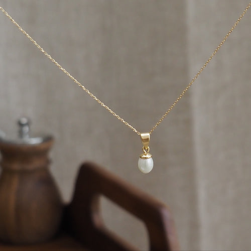 A delicate gold chain necklace with a small gold cap and a single natural pearl pendant, displayed against a soft beige linen background with a blurred wooden object in the foreground.
