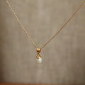 A delicate gold chain necklace with a small white pearl pendant, displayed against a soft-focus beige background.