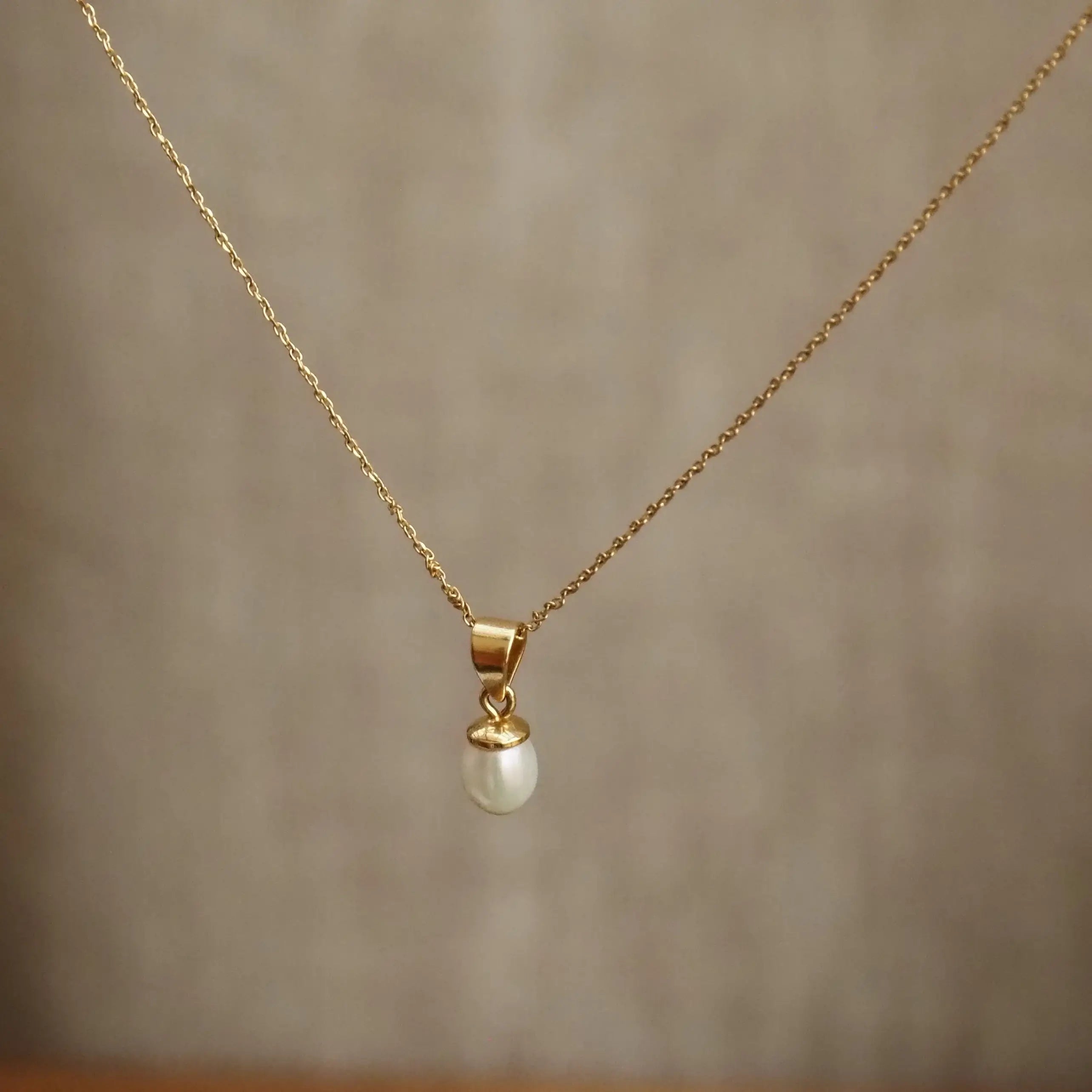 A delicate gold chain necklace with a small white pearl pendant, displayed against a soft-focus beige background.