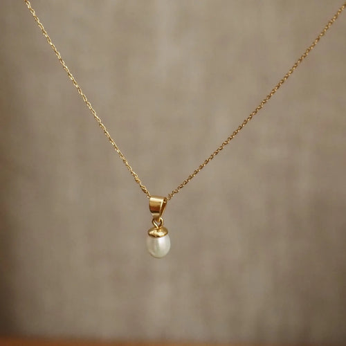 A delicate gold chain necklace with a small white pearl pendant, displayed against a soft-focus beige background.