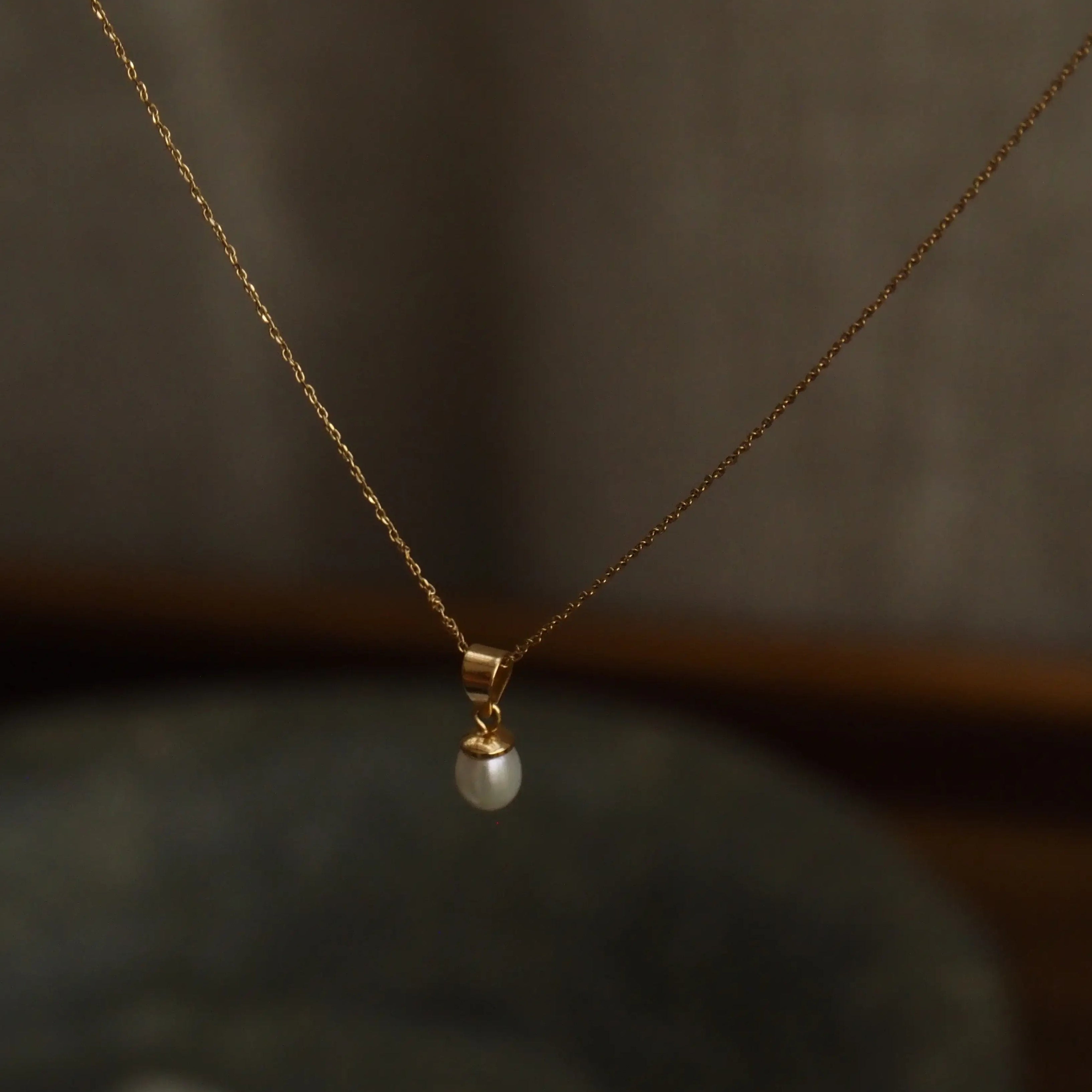 A delicate gold chain necklace with a small white pearl pendant, suspended against a dark, softly blurred background.