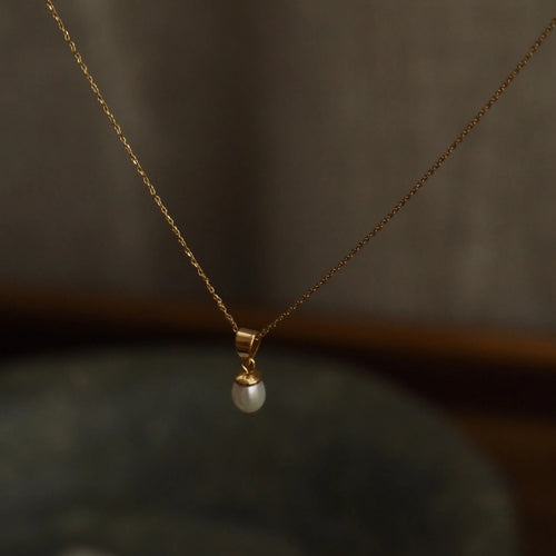A delicate gold chain necklace with a small white pearl pendant, suspended against a dark, softly blurred background.