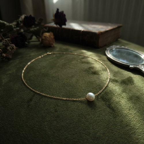 Gold necklace with a pearl pendant on a green surface with dried flowers and a book in the background.