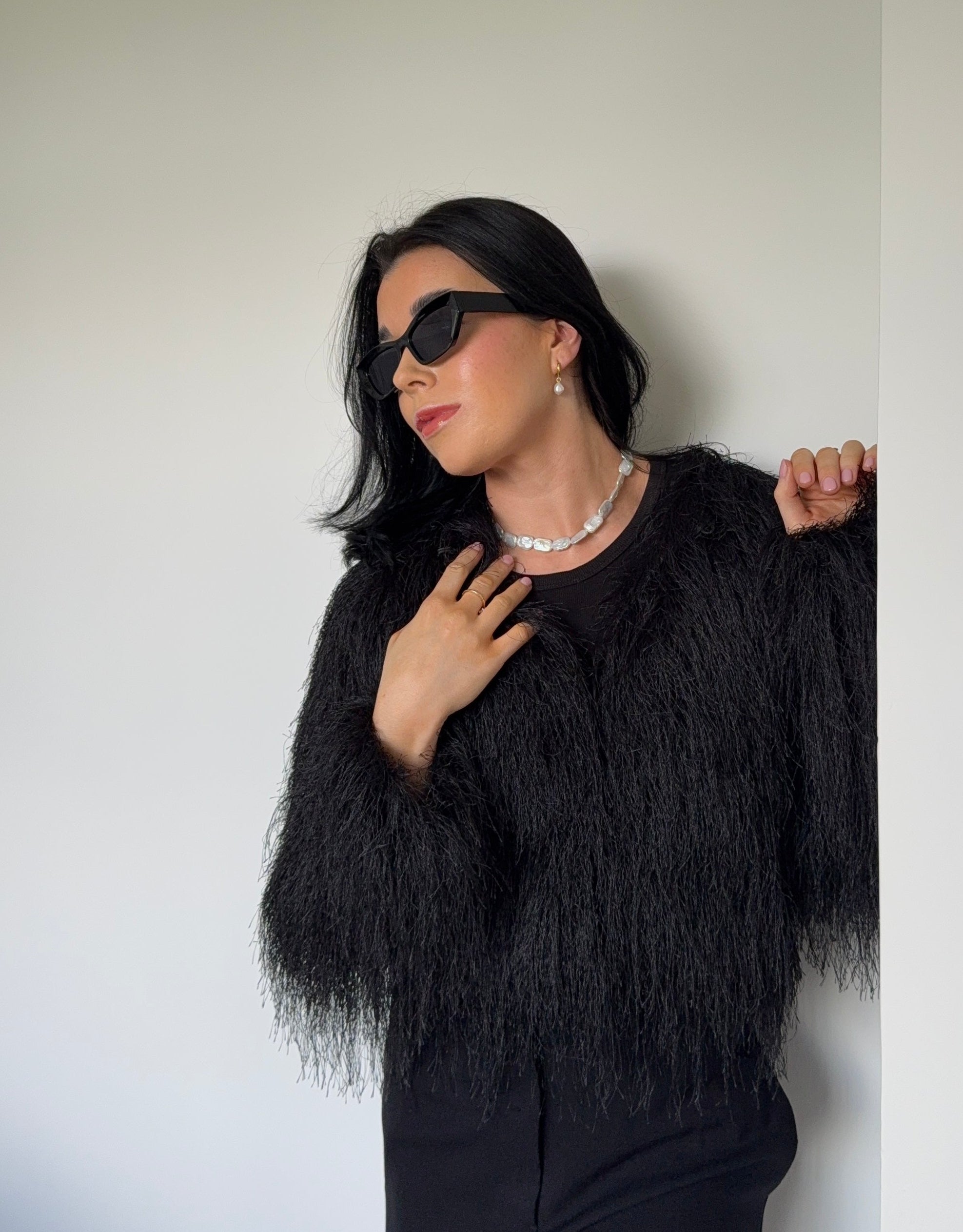 Woman wearing a black textured sweater and sunglasses against a plain wall.