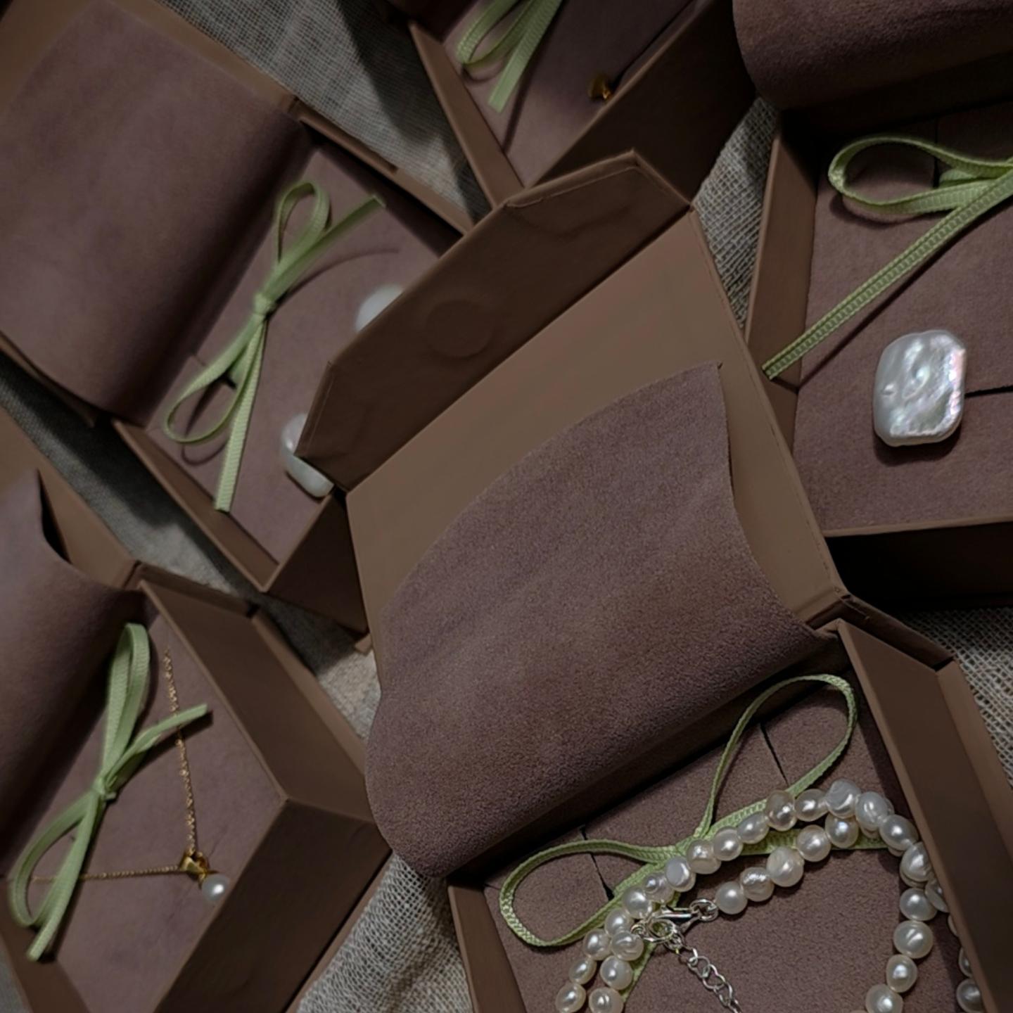 Several brown jewelry gift boxes arranged on a textured fabric surface, each containing pearl necklaces and a white shell pendant tied with pale green ribbon.