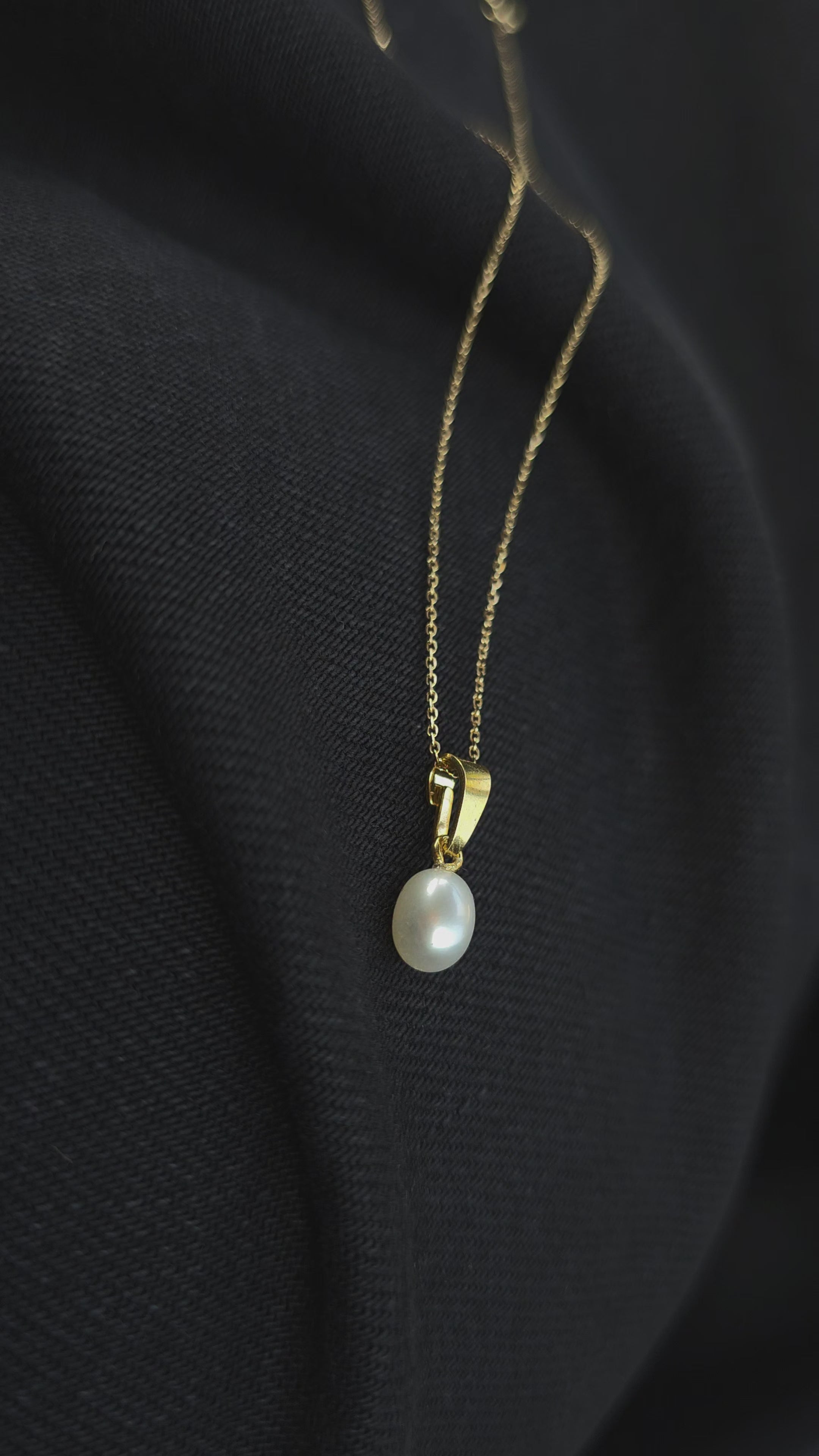 Close‑up of a gold‑plated silver necklace with a single oval freshwater pearl pendant against dark fabric.
