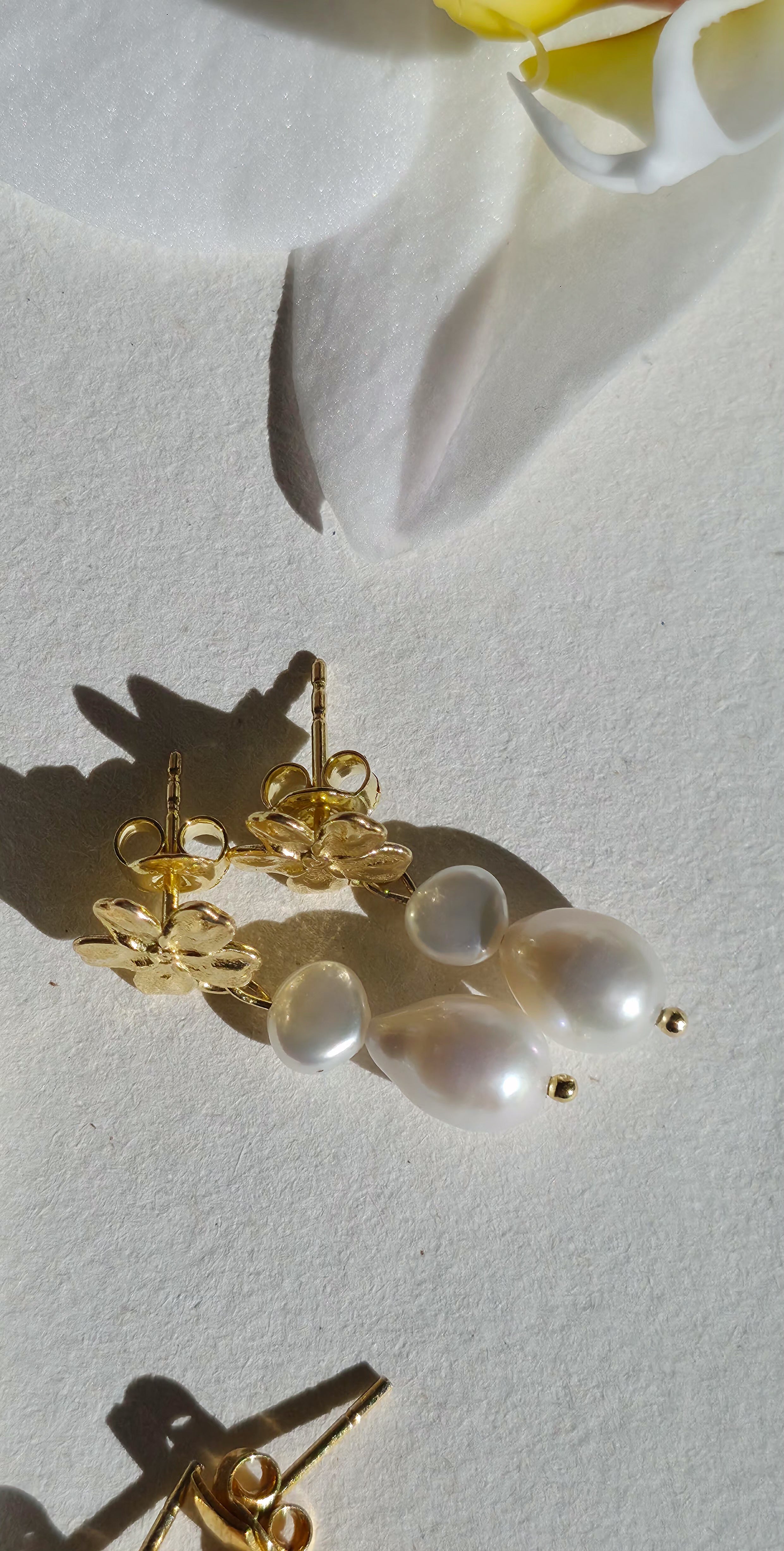 Gold earrings with pearl‑like beads styled beside a white flower on a light background.