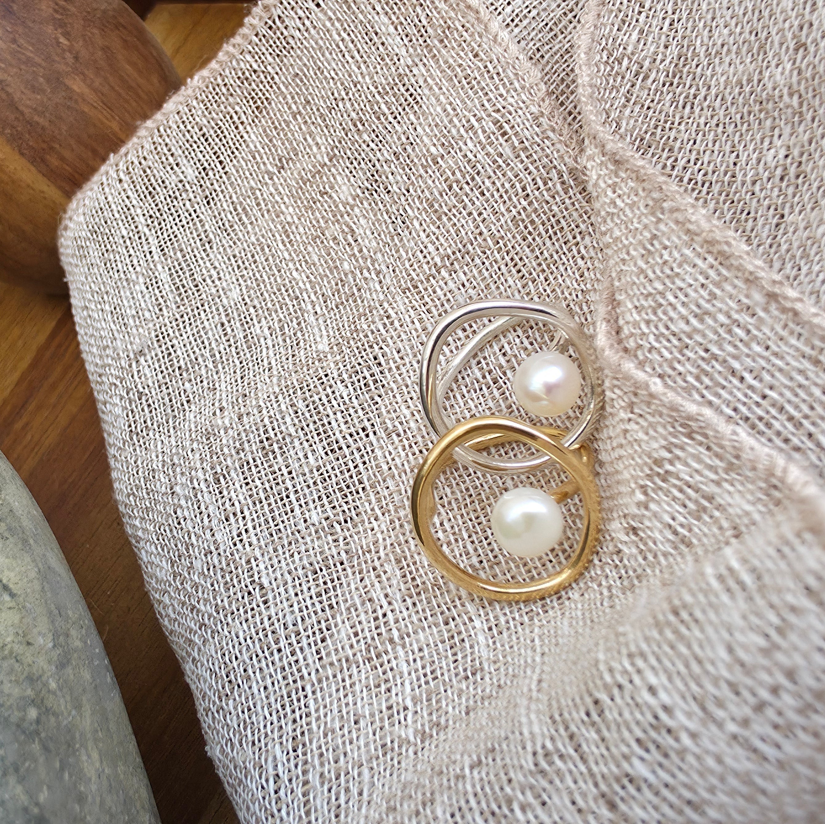 A pair of gold and silver rings with a floating pearl, displayed on a textured beige linen fabric.