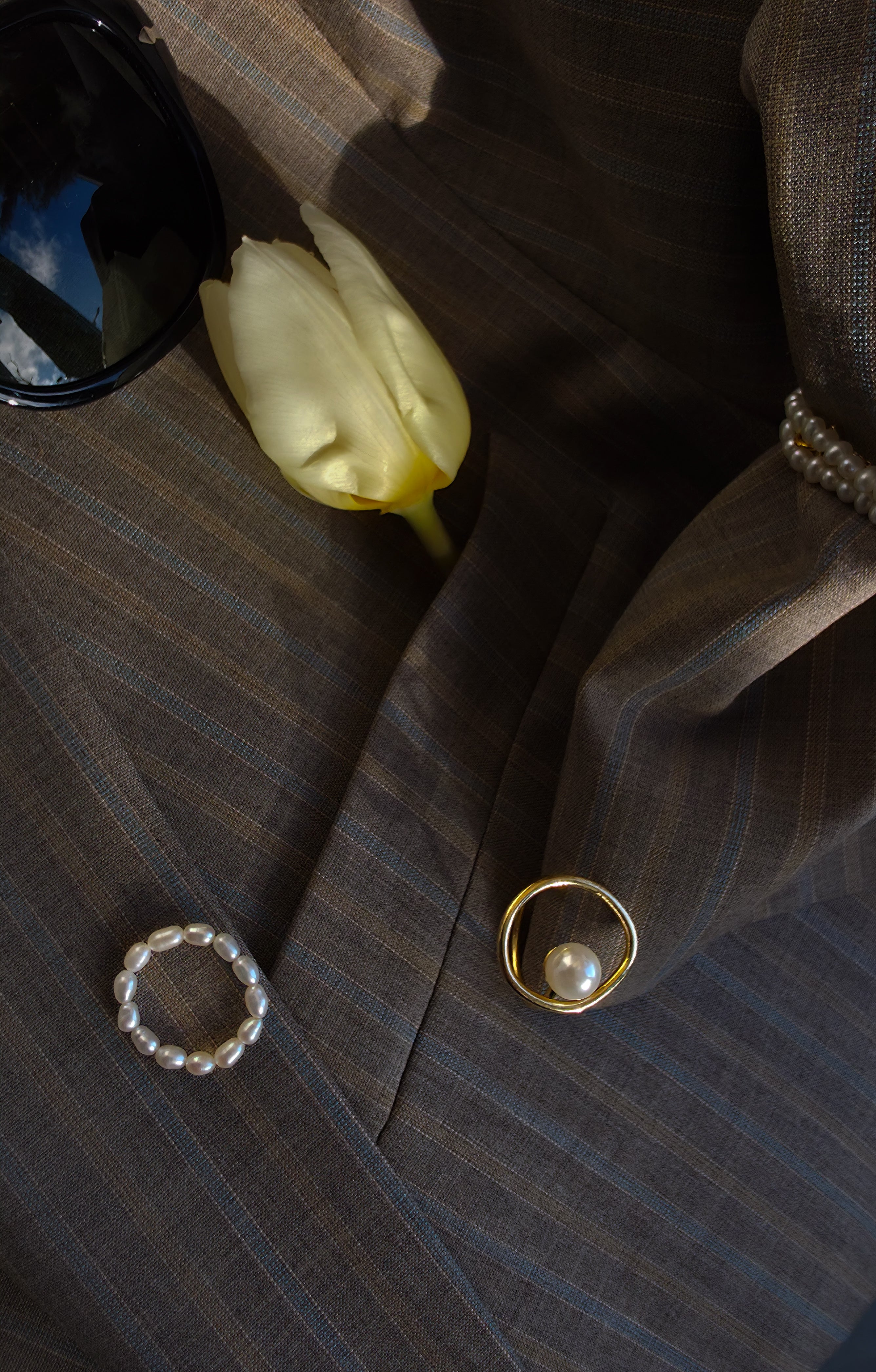 Close-up of a suit jacket, a pearl ring layered on, a yellow tulip, and a black pair of sunglasses on a dark surface.