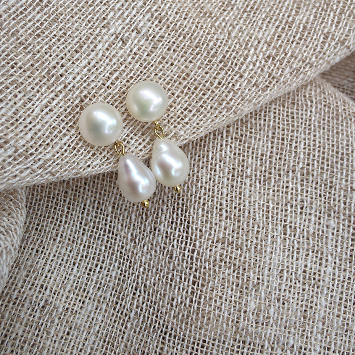 Three baroque pearl stud earrings with gold-tone settings resting on a textured beige linen fabric.