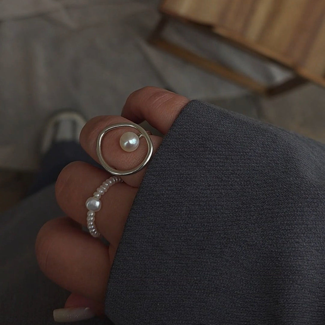 A close-up of a hand wearing two silver rings, one with a large oval bezel set with a white pearl and the other a delicate pearl-studded band, against a gray fabric background.
