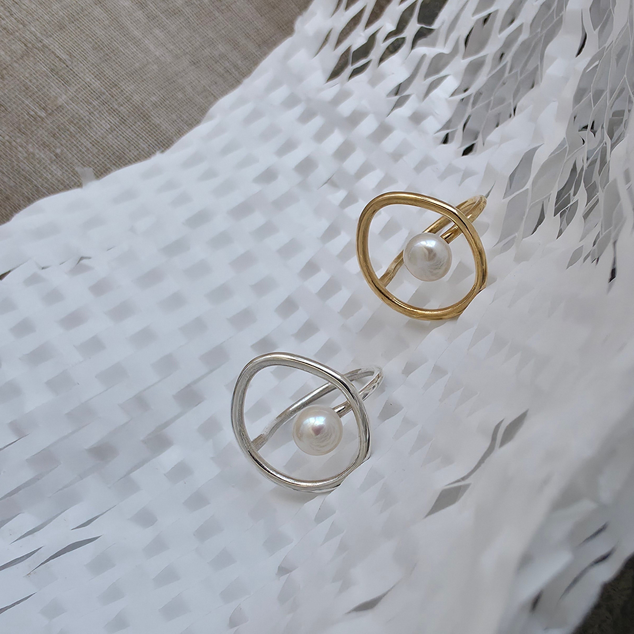 Two minimalist open silhouettes rings with pearl centers, one in gold-tone silver and one in silver metal, displayed on a white woven textile.