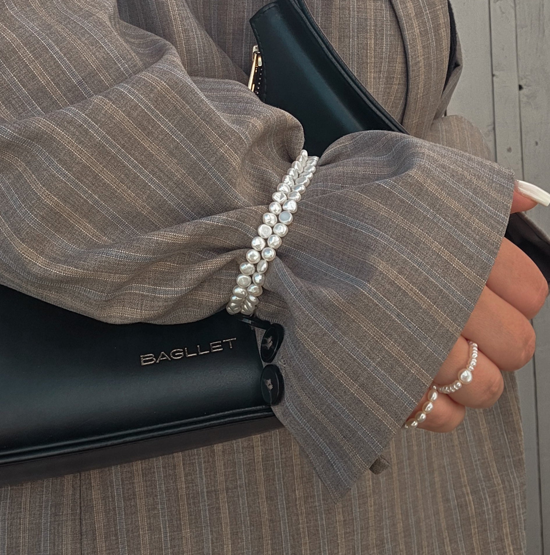 Person holding a black handbag with 'Luca Di Raffaello' branding, wearing pearl rings and a gray pinstripe suit.