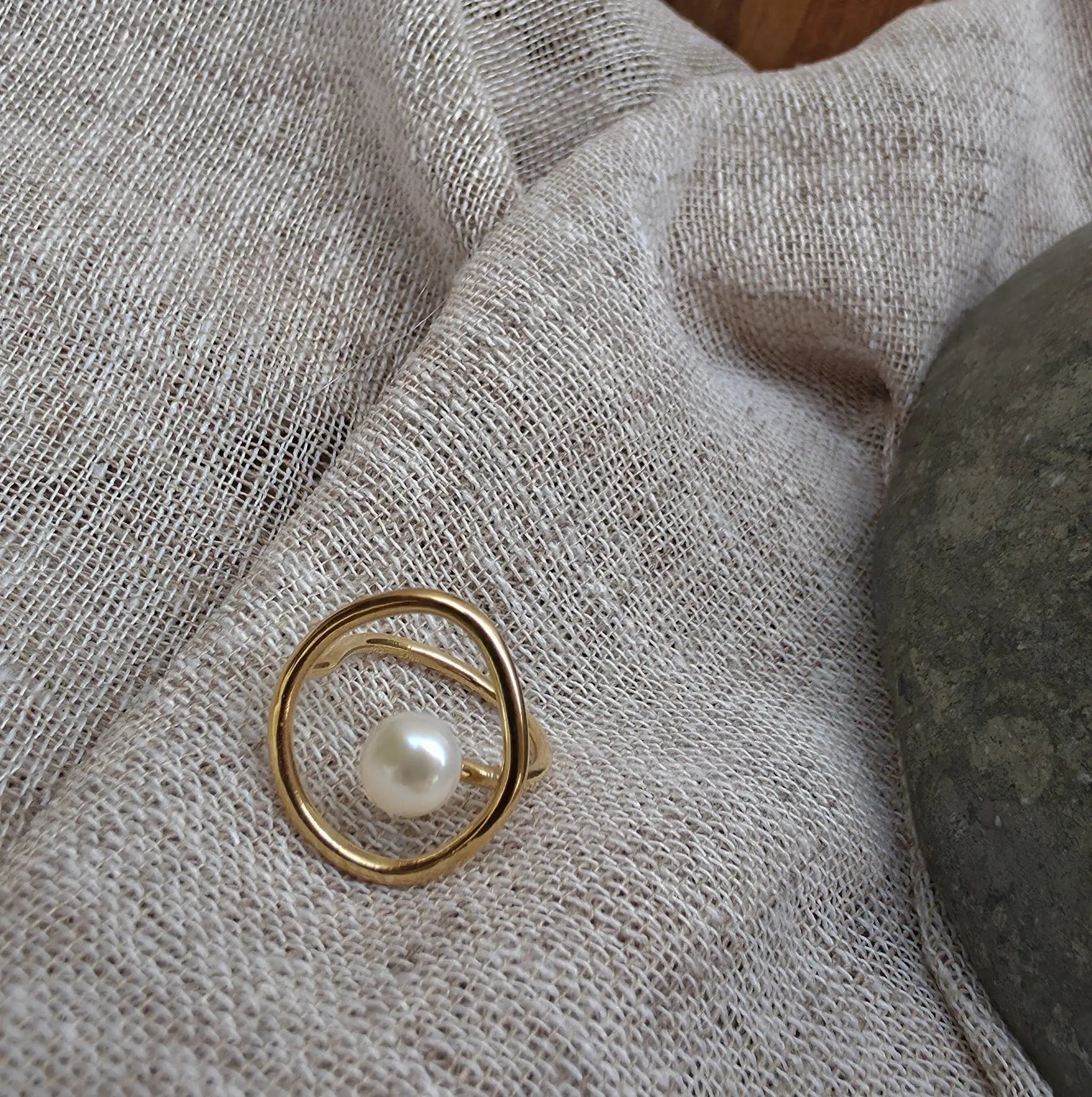 A gold ring with a single white pearl, resting on a textured beige linen fabric next to a dark gray stone.
