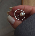 A silver ring with a single pearl, held between fingers. The ring features an asymmetrical design with a flat, wide oval band on one side and a thin band on the other holding the pearl.