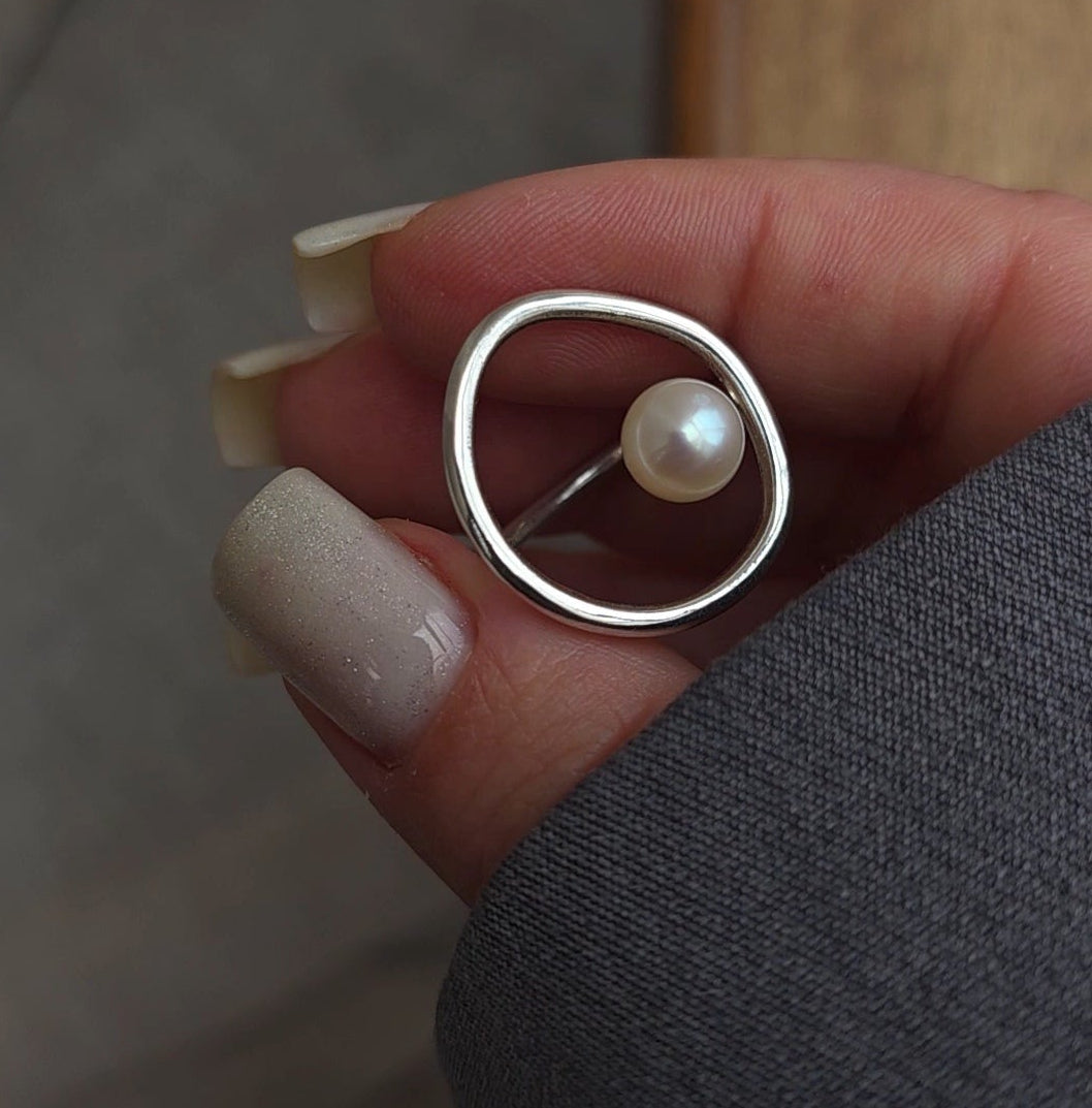 A silver ring with a single pearl, held between fingers. The ring features an asymmetrical design with a flat, wide oval band on one side and a thin band on the other holding the pearl.