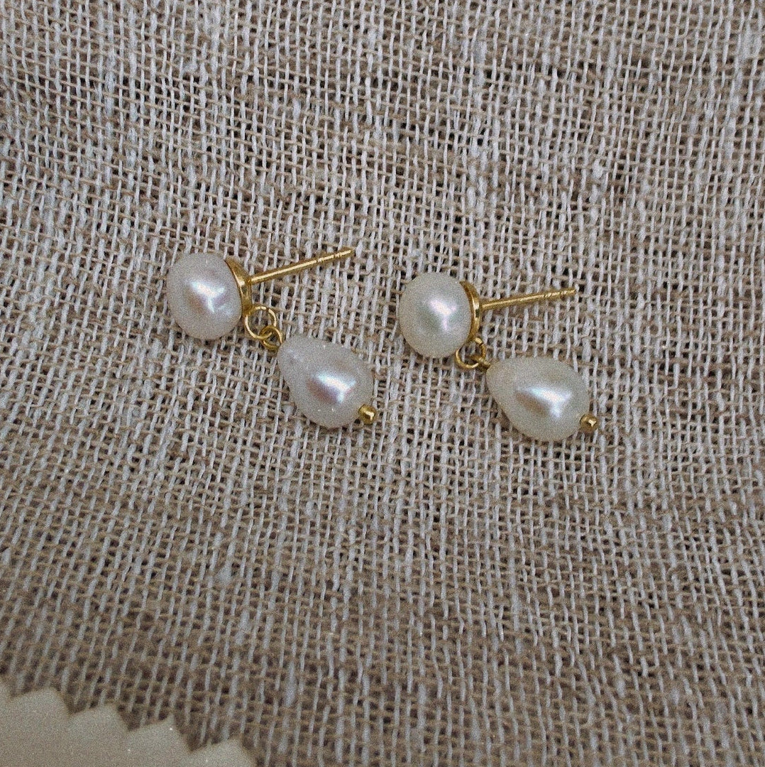 A pair of gold stud earrings with irregular white freshwater pearls, displayed on a textured beige linen fabric background.