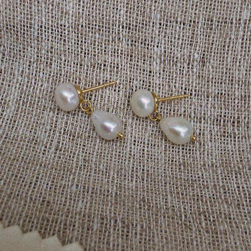 A pair of gold stud earrings with irregular white freshwater pearls, displayed on a textured beige linen fabric background.