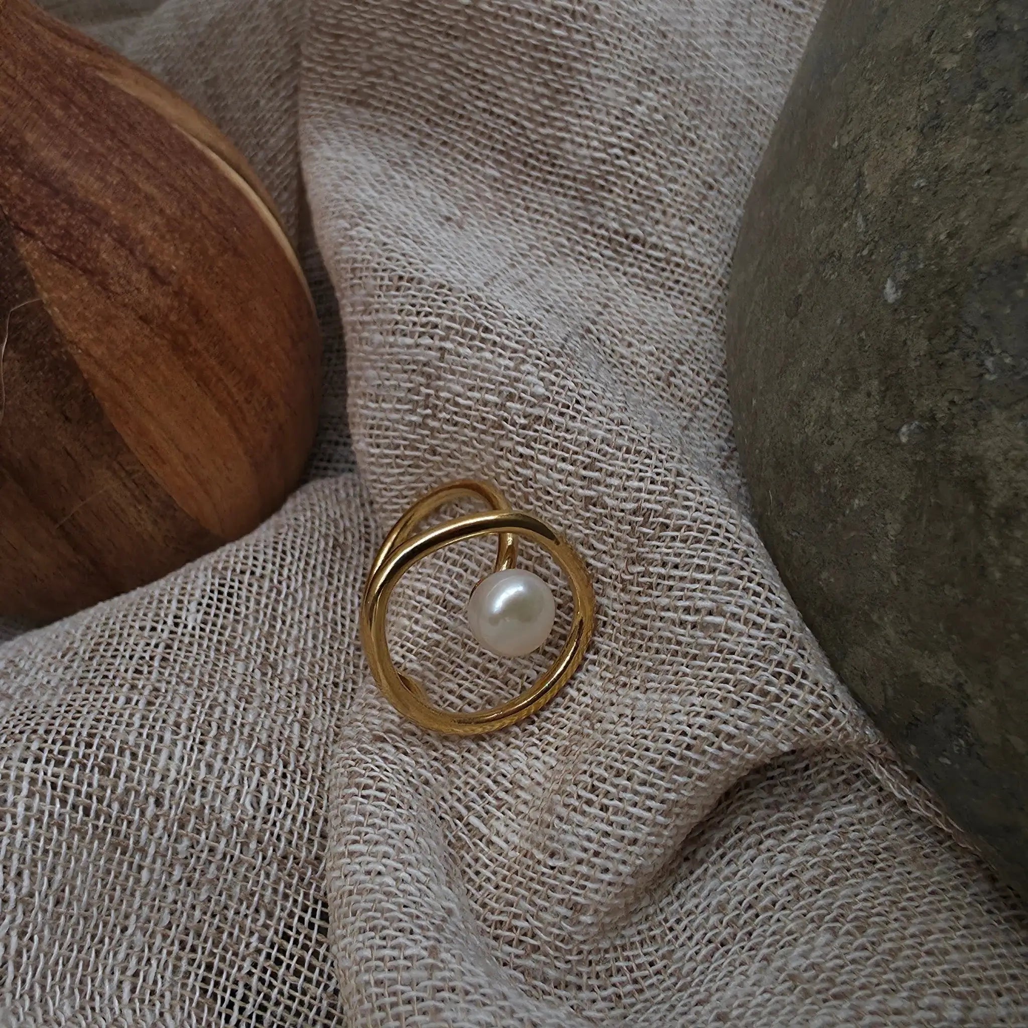 A gold ring with a single white pearl set in a circular open design, resting on a textured beige linen fabric with a wooden spoon and a dark stone prop.