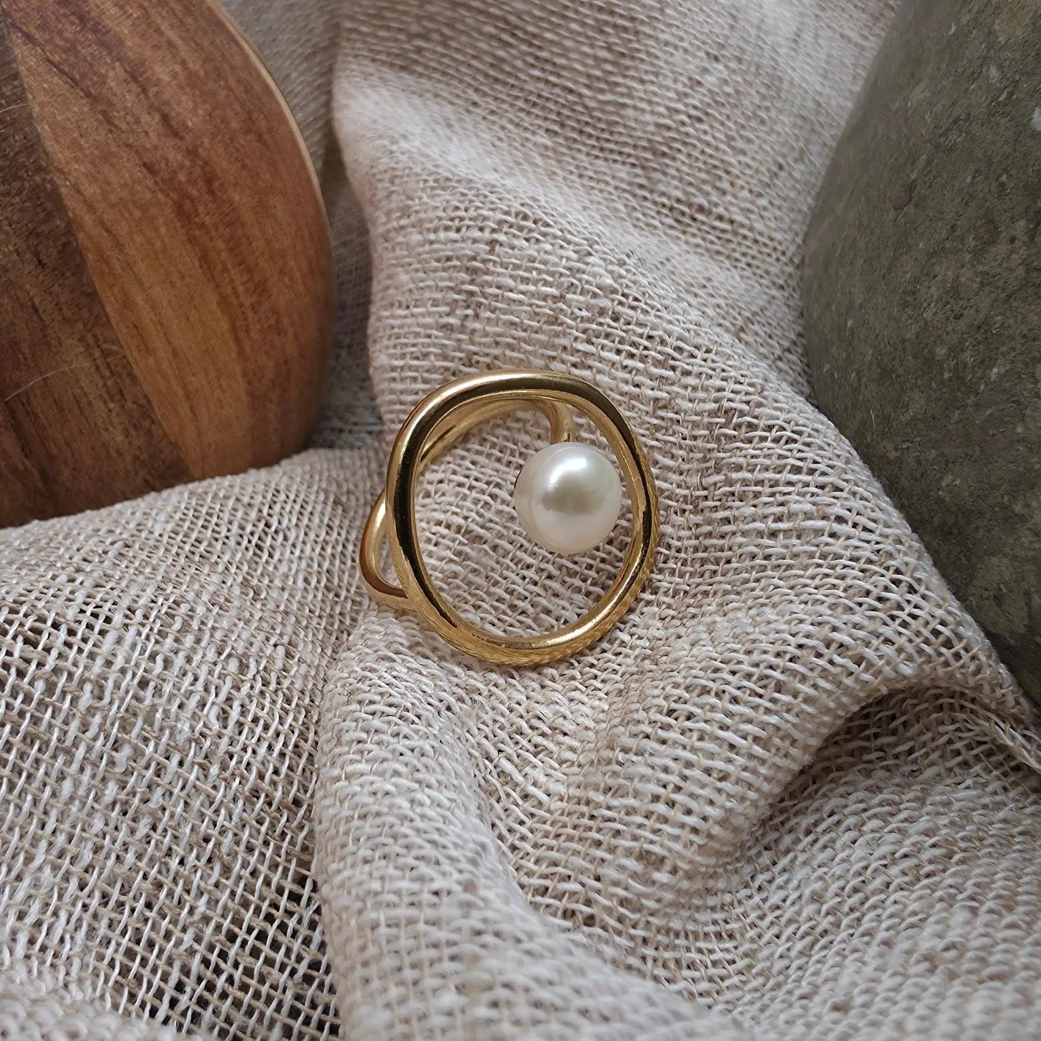 A gold ring with a single white pearl set in a circular, open design, resting on a textured beige linen fabric.